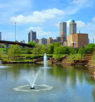 Centennial Park in Tulsa, Oklahoma with a water fountain in the middle of a lake in front of a bridge and city buildings.
