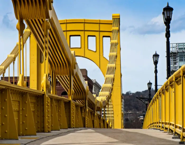 Yellow Andy Warhol bridge in Pittsburgh leading into a city with lamp posts in a row on the side.