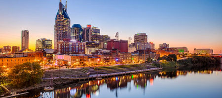 City skyline of Nashville, Tennessee at dawn over a body of water with buildings reflected and lit up with warm lights.