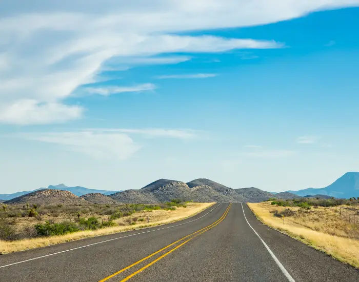 West Texas roads in the summer with small mountains, yellow grass, and a partly cloudy blue sky.