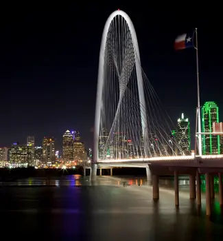 Dallas, Texas bridge illuminated at night with lights reflecting off the waters and a state flag flying.