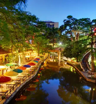 Vibrant River Walk in San Antonio at night with colored umbrellas, overgrown trees, and shopping centers.