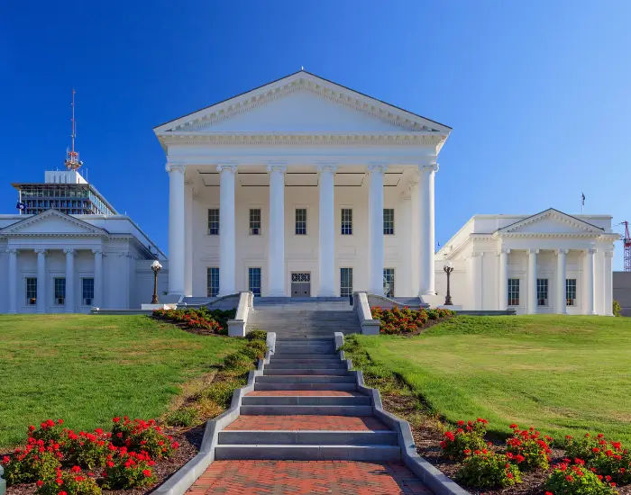 White Virginia State Capitol building in Richmond, Virginia with a red brick path leading up staircases and red flowers surrounding.
