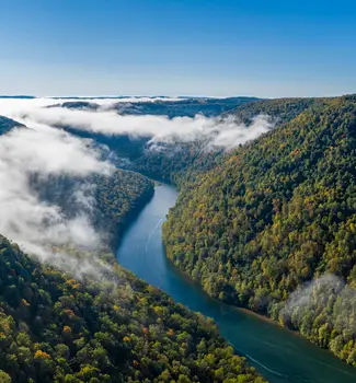 Aerial drone footage of a river flowing through Cheat River Canyon in West Virginia with clouds, clear blue sky, and thick forest trees surrounding the water.