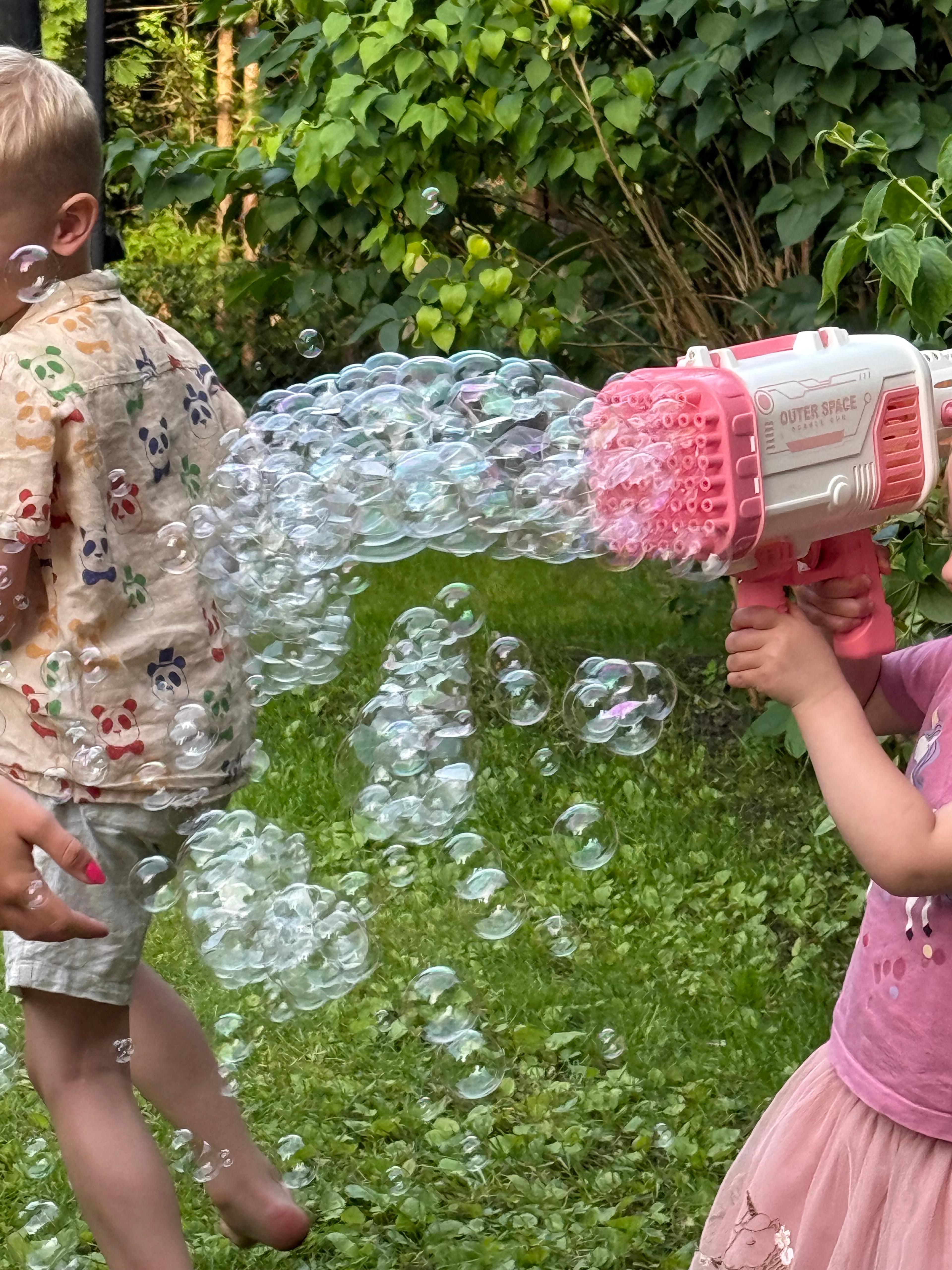 A boy and a girl playing in a green garden with a large pink and white bubble gun. The girl is aiming the gun, and a massive cloud of connected bubbles streams towards the boy, who is running away.