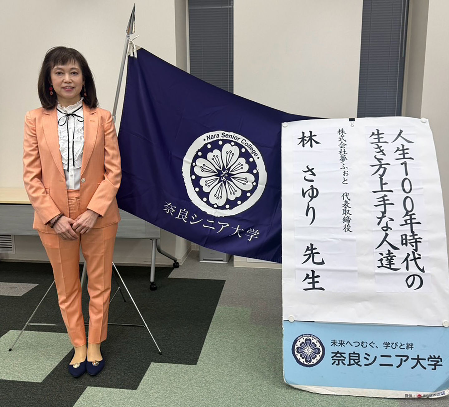 奈良シニア大学の大学旗と講座看板の前に立つ林さゆり