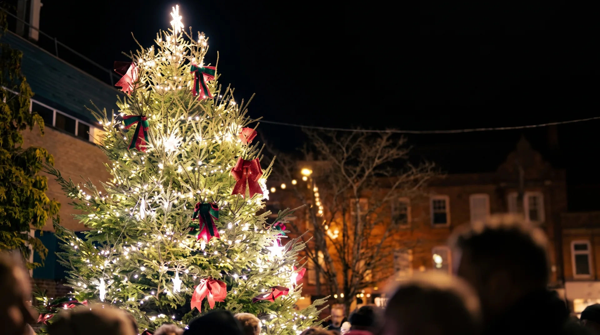 A beautifully lit Christmas tree adorned with red and green ribbons at night, surrounded by a festive crowd.