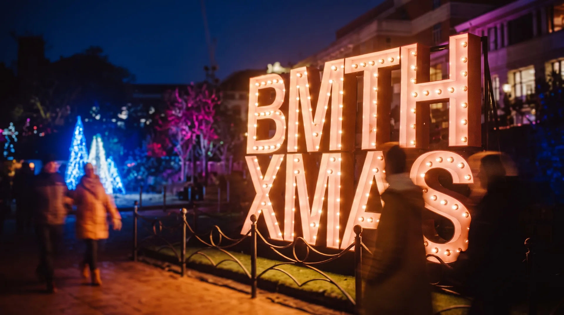 Brightly lit 'BMTH XMAS' sign with festive trees in the background at night