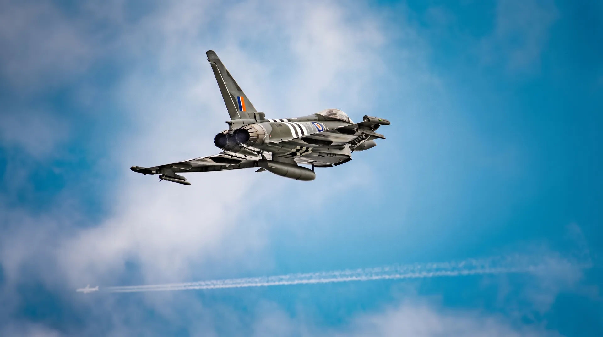A military jet flying against a blue sky with wispy clouds and a faint contrail in the distance