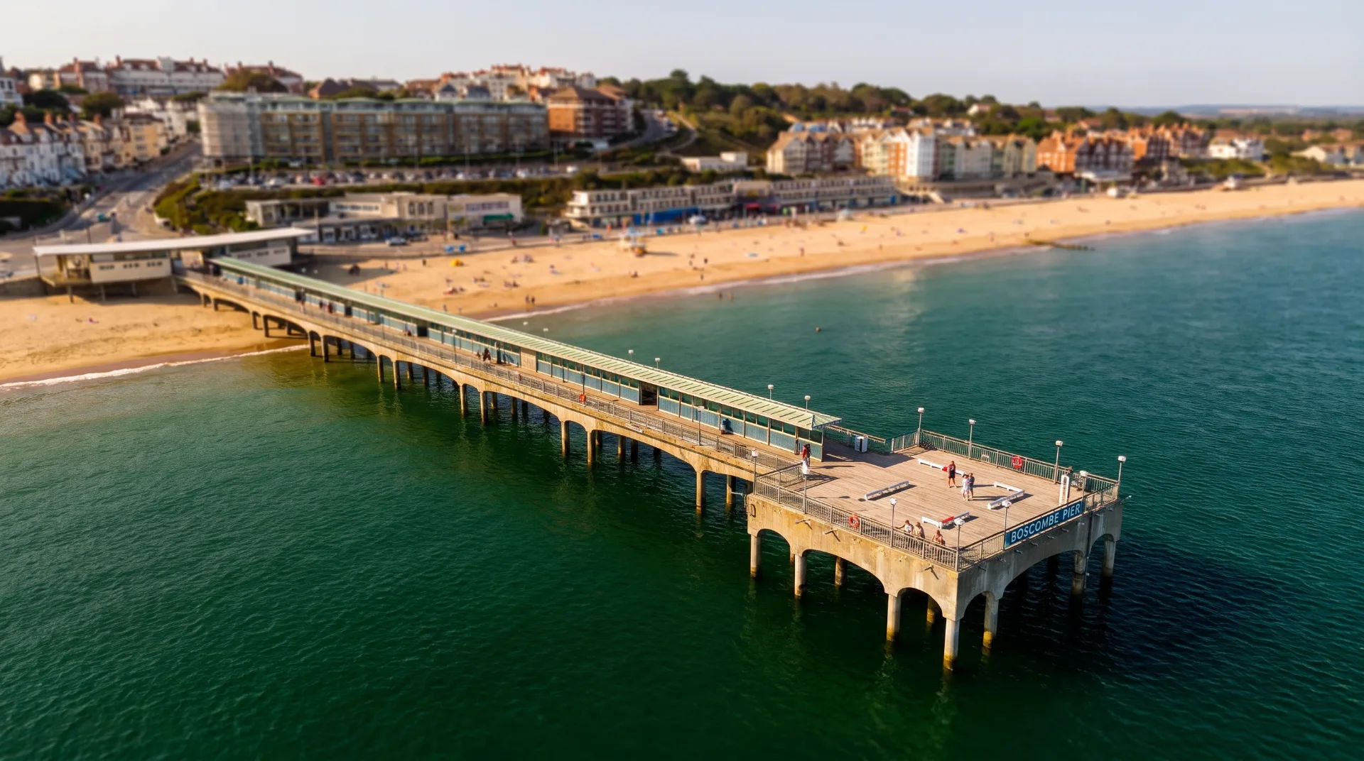 Aerial view of Boscombe Pier extending over calm waters, with sandy beach and colourful buildings in the background.