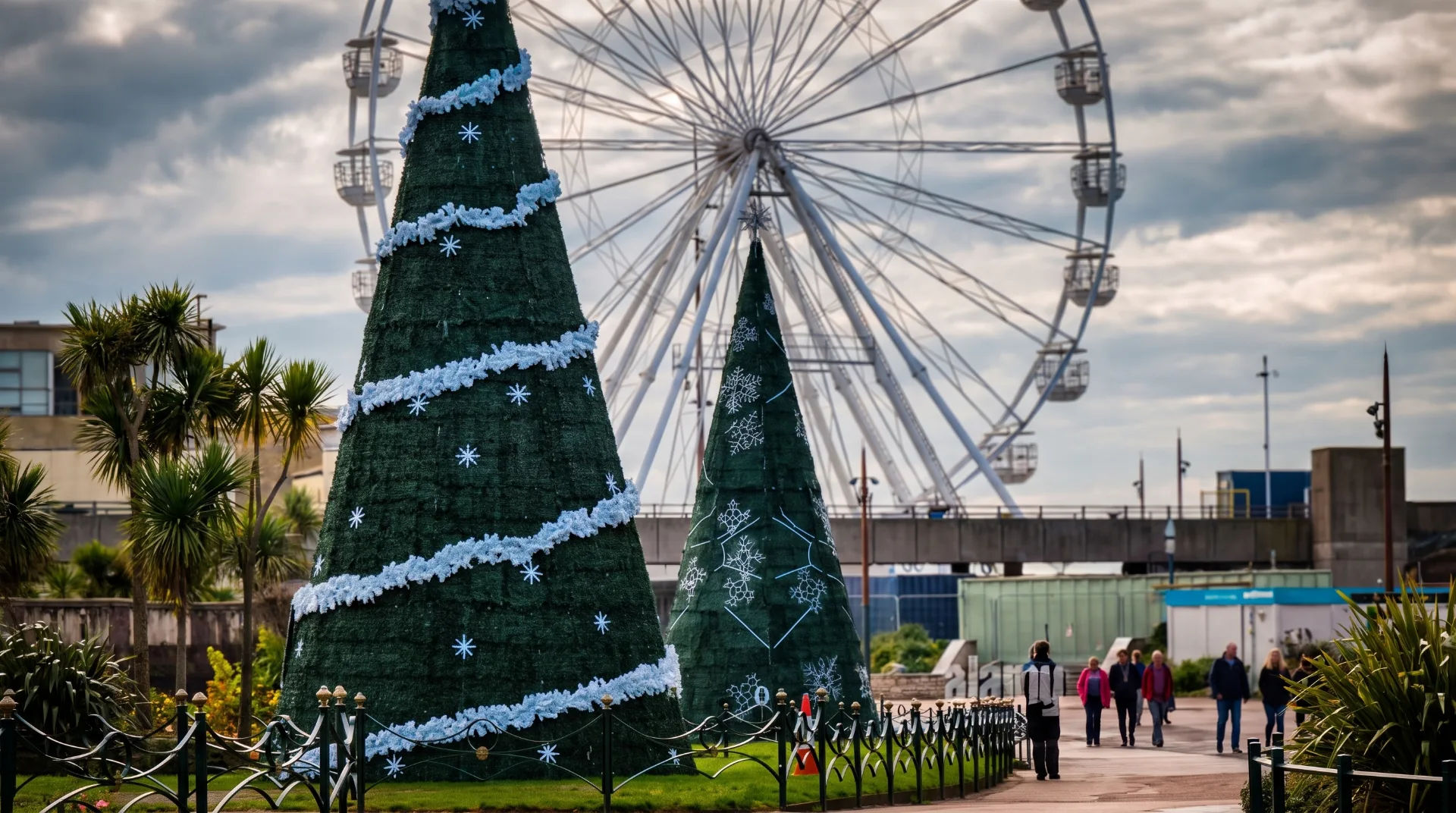 Two large decorated Christmas trees in a park with a Ferris wheel in the background and people walking nearby.