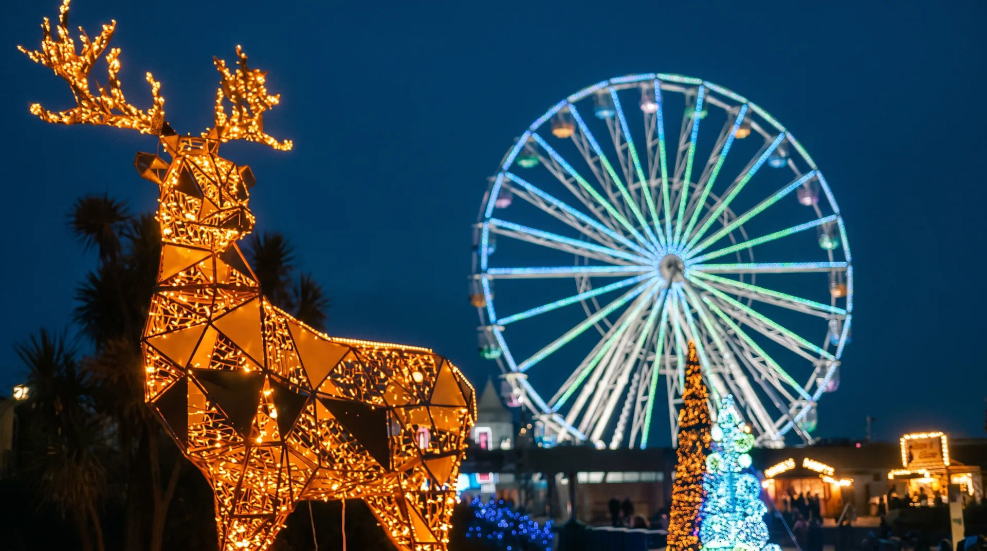 Illuminated reindeer sculpture with a ferris wheel in the background, decorated for Christmas at night.