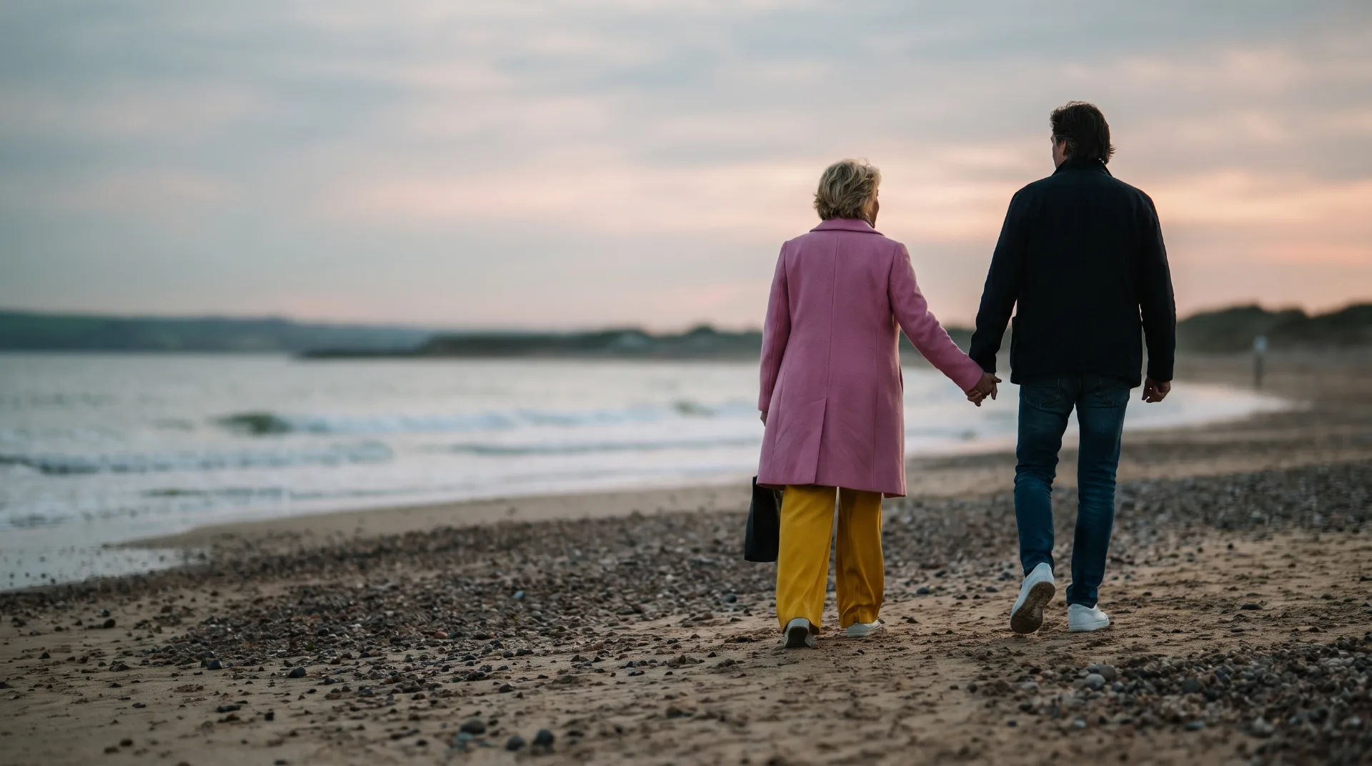 A couple walking hand in hand along a pebbled beach at sunset, with gentle waves lapping at the shore.