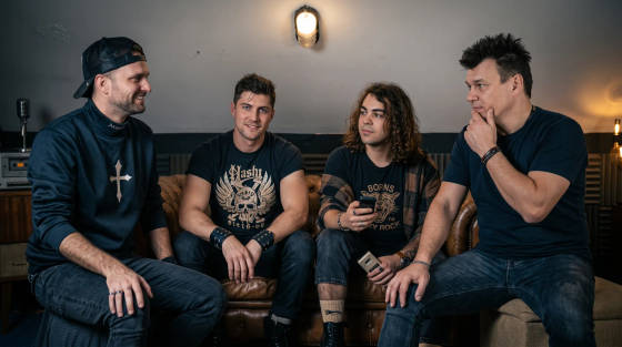 Four men in casual clothing sitting on a couch in a music studio