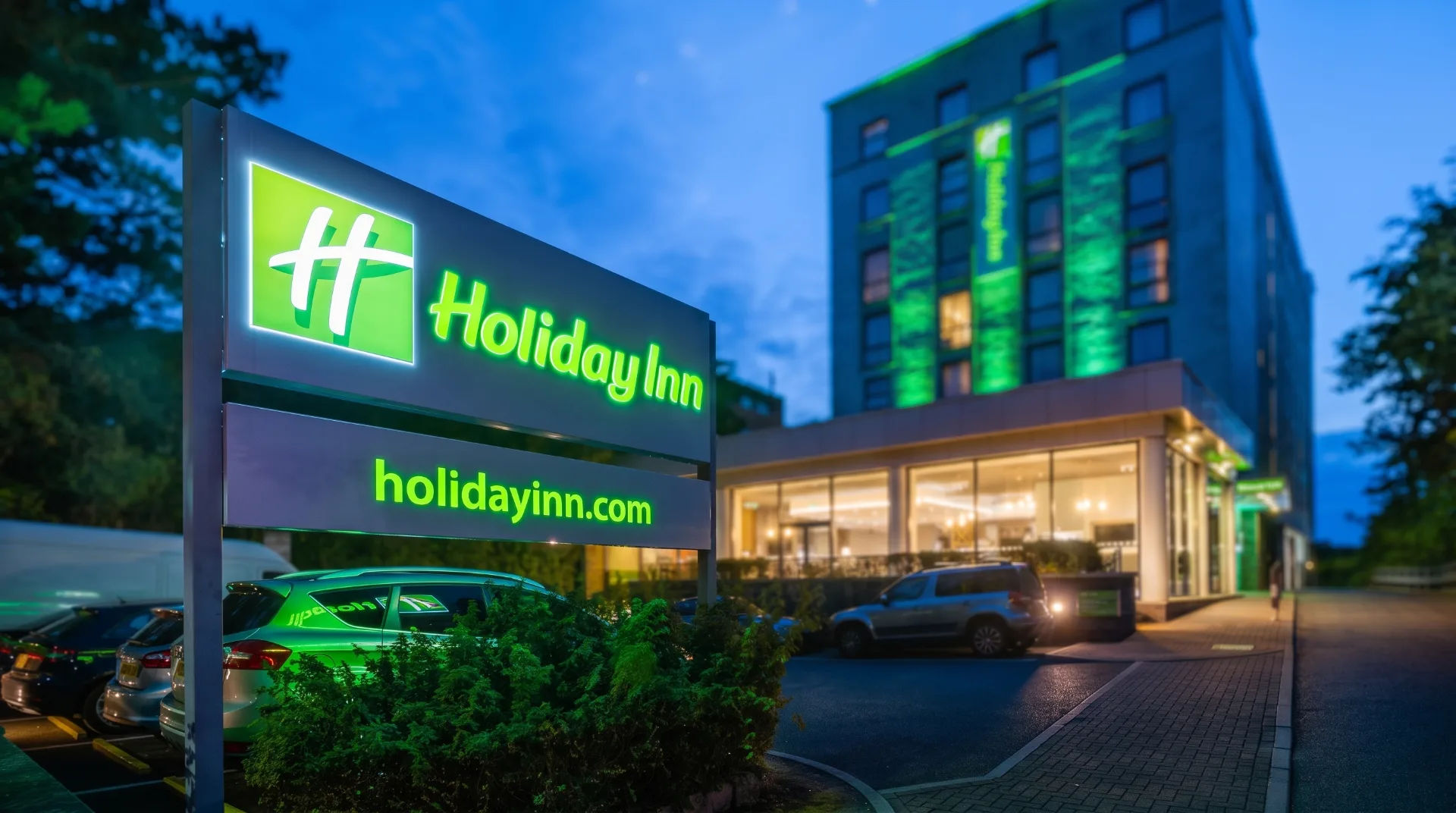 Holiday Inn sign illuminated at night with hotel entrance and parked cars visible