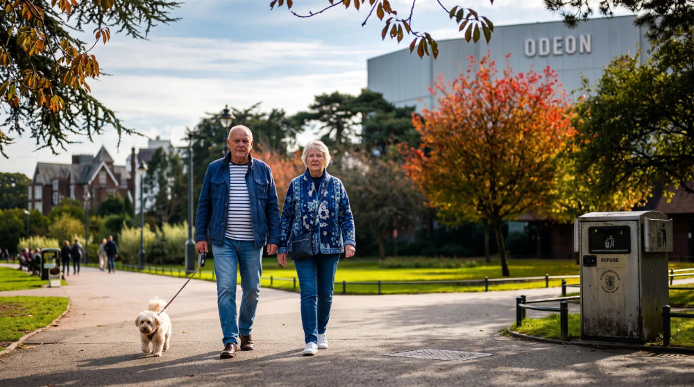 An older couple walking a dog along a path with autumn trees and the Odeon cinema in the background.