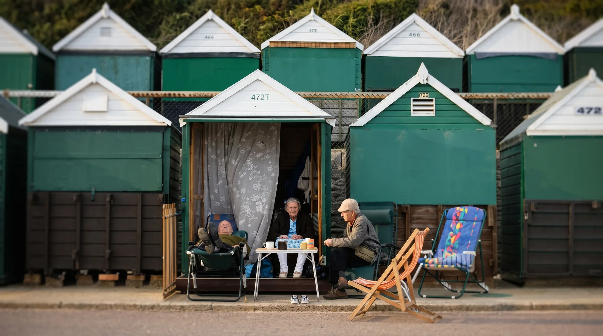 Three people relax outside beach huts, enjoying drinks and conversation on a sunny day.