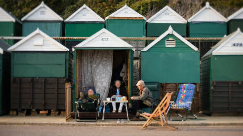 Three people sitting outside beach huts with green and white roofs