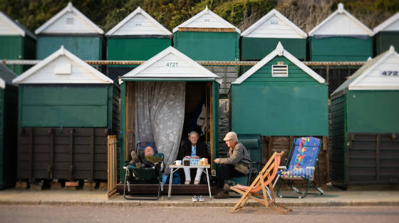 Three people sitting outside beach huts with green and white roofs