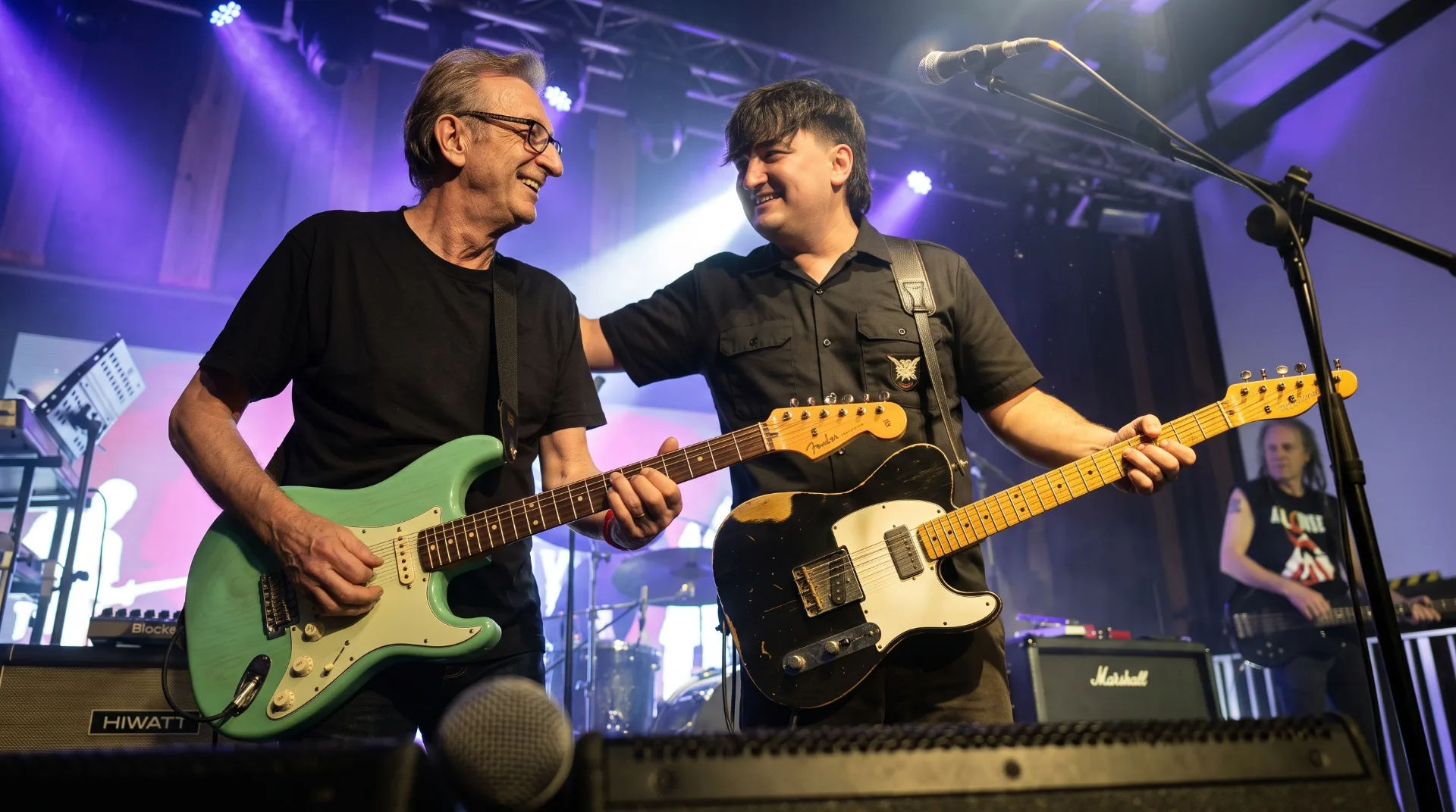 Two guitarists share a moment on stage, one with a mint green guitar and the other with a black Telecaster.