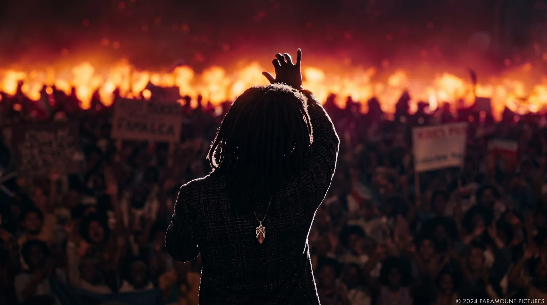 Silhouette of a performer with dreadlocks raising a hand in front of a large crowd and a fiery background