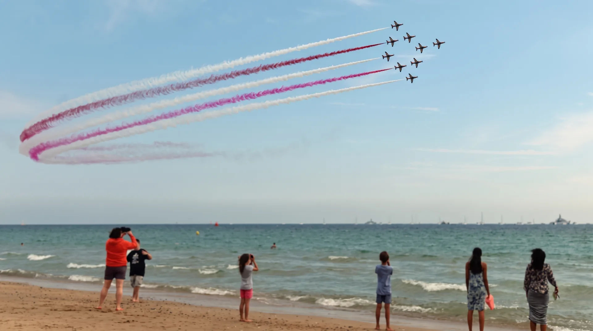 People on the beach watching an aerial display with planes leaving colourful smoke trails in the sky.