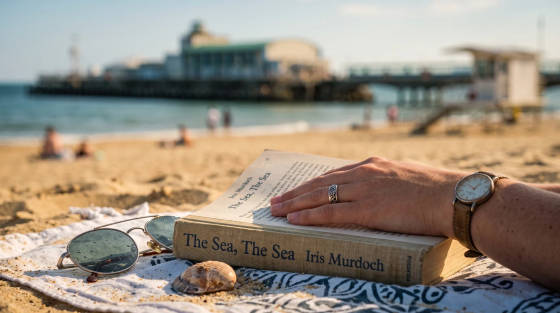 A hand resting on a book on the sand with sunglasses nearby