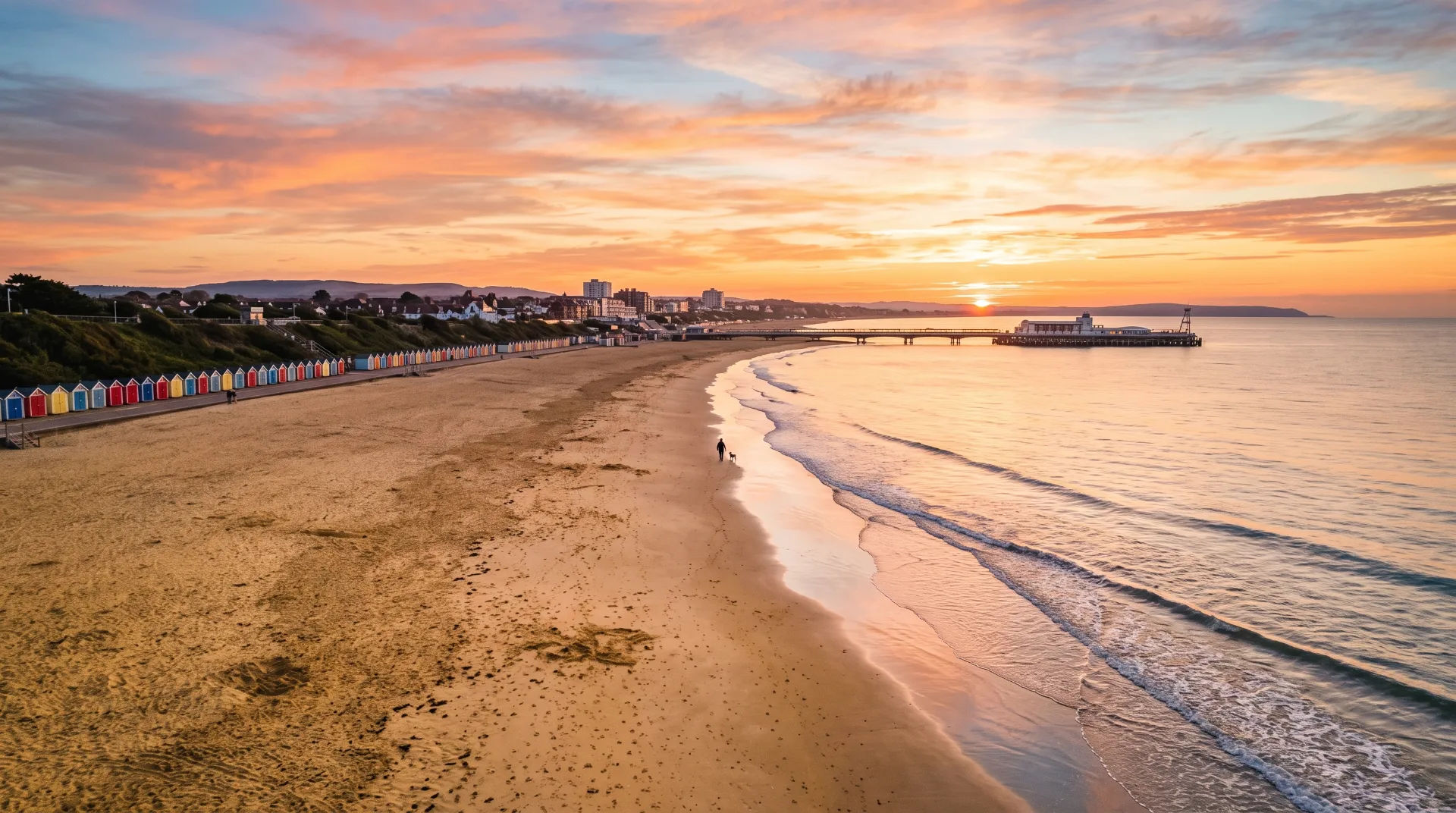 A sandy beach with colourful beach huts, a person walking a dog, and a pier at sunset.