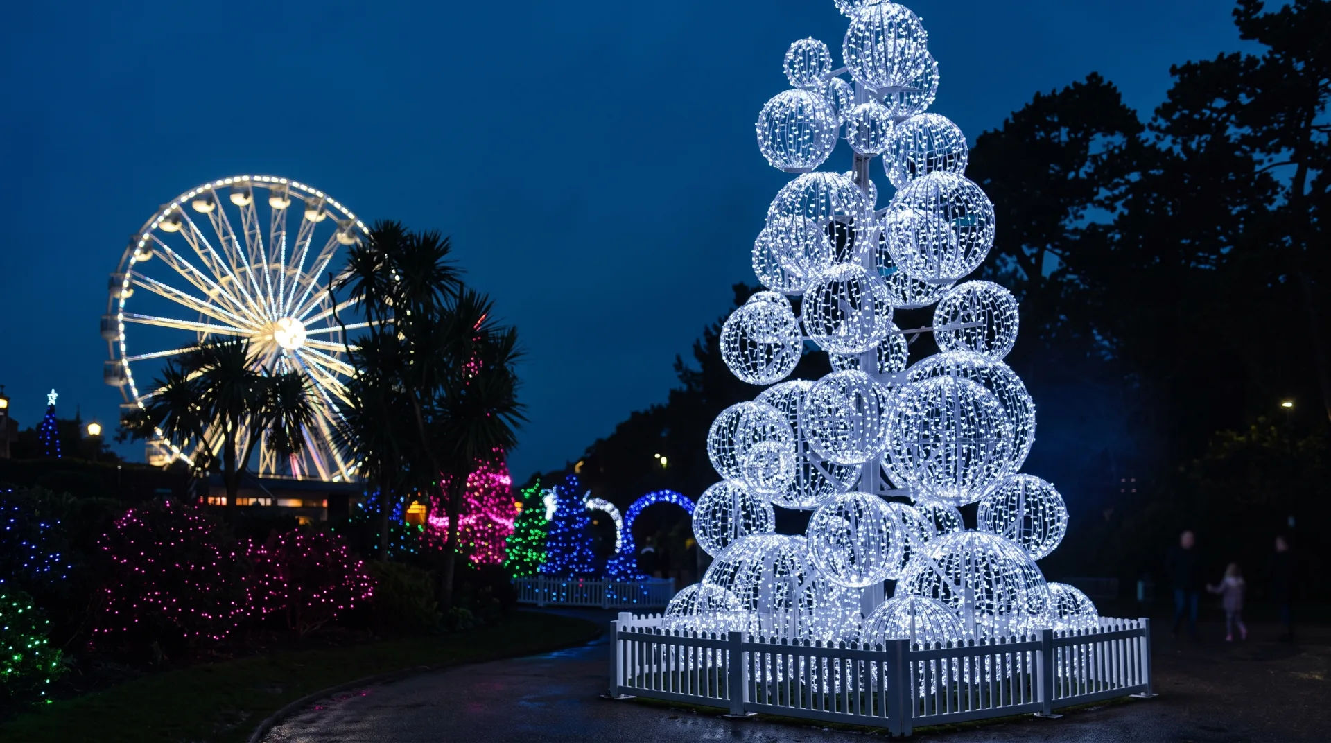 A large illuminated Christmas tree made of white lights, with a Ferris wheel and colourful lights in the background.