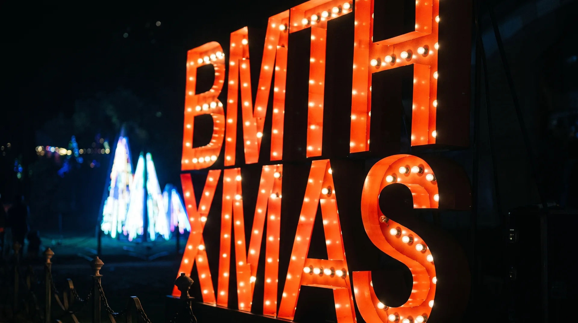 Brightly lit sign reading 'BMTH XMAS' against a dark background with illuminated trees nearby