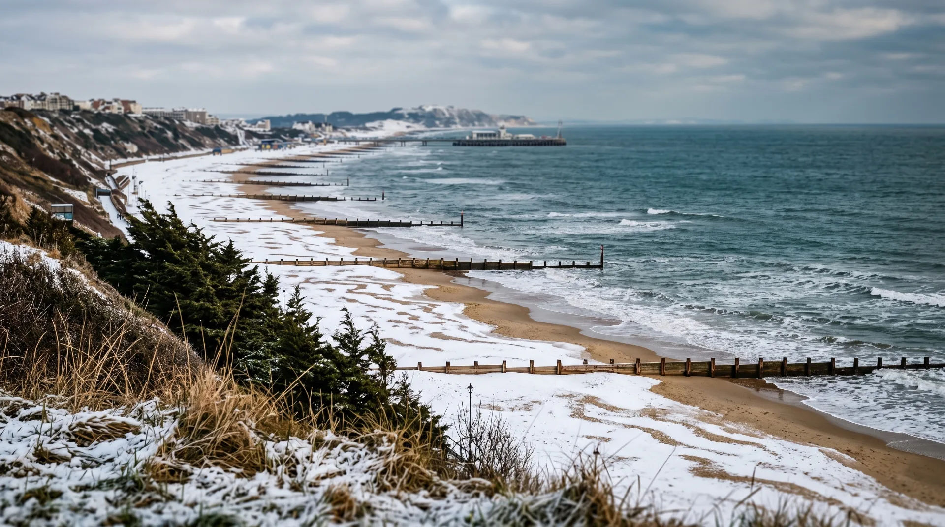 Snow-covered beach with wooden groynes and waves, under a cloudy sky