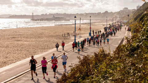 A group of runners on a promenade with the beach and pier in the background
