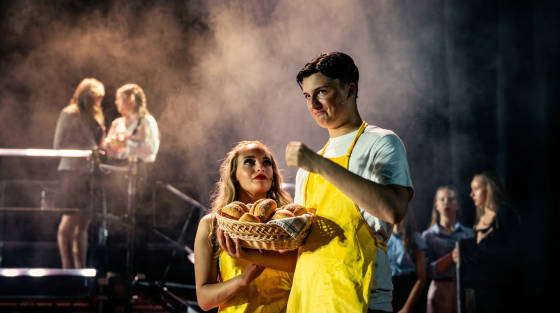 Two actors on stage holding a basket of bread rolls in a smoky atmosphere