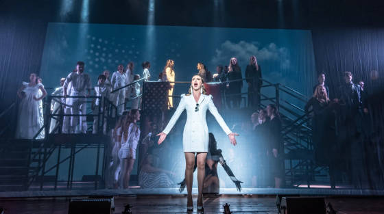 A singer in a white dress performing on stage with a choir behind