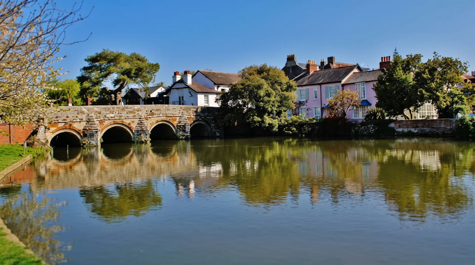 Stone bridge with arches over calm water, surrounded by trees and quaint buildings in pastel colours