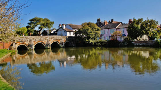 A stone bridge with arches over a calm river surrounded by trees and houses
