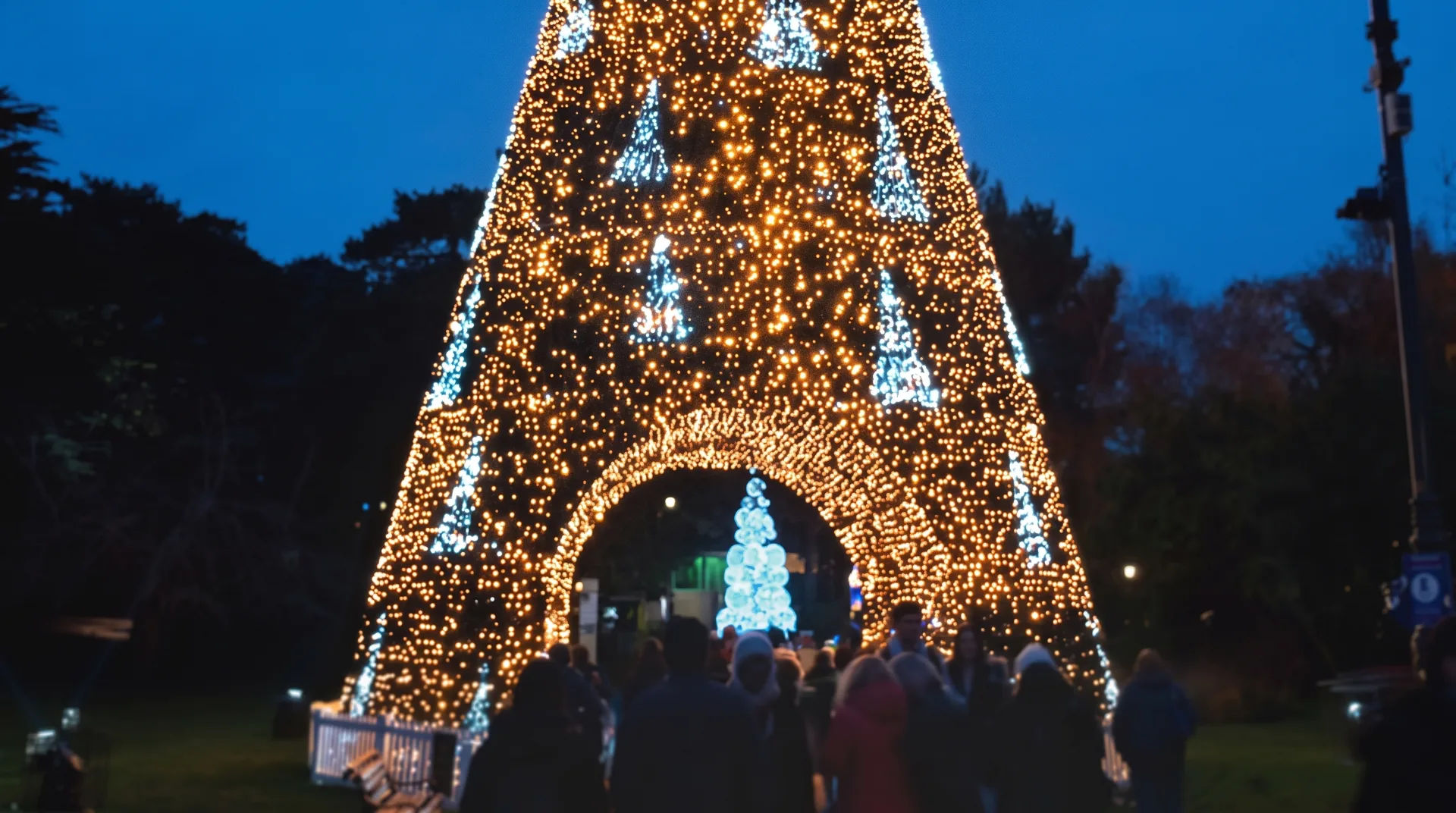 A large illuminated Christmas tree structure with blue and white lights and people walking underneath it at dusk.