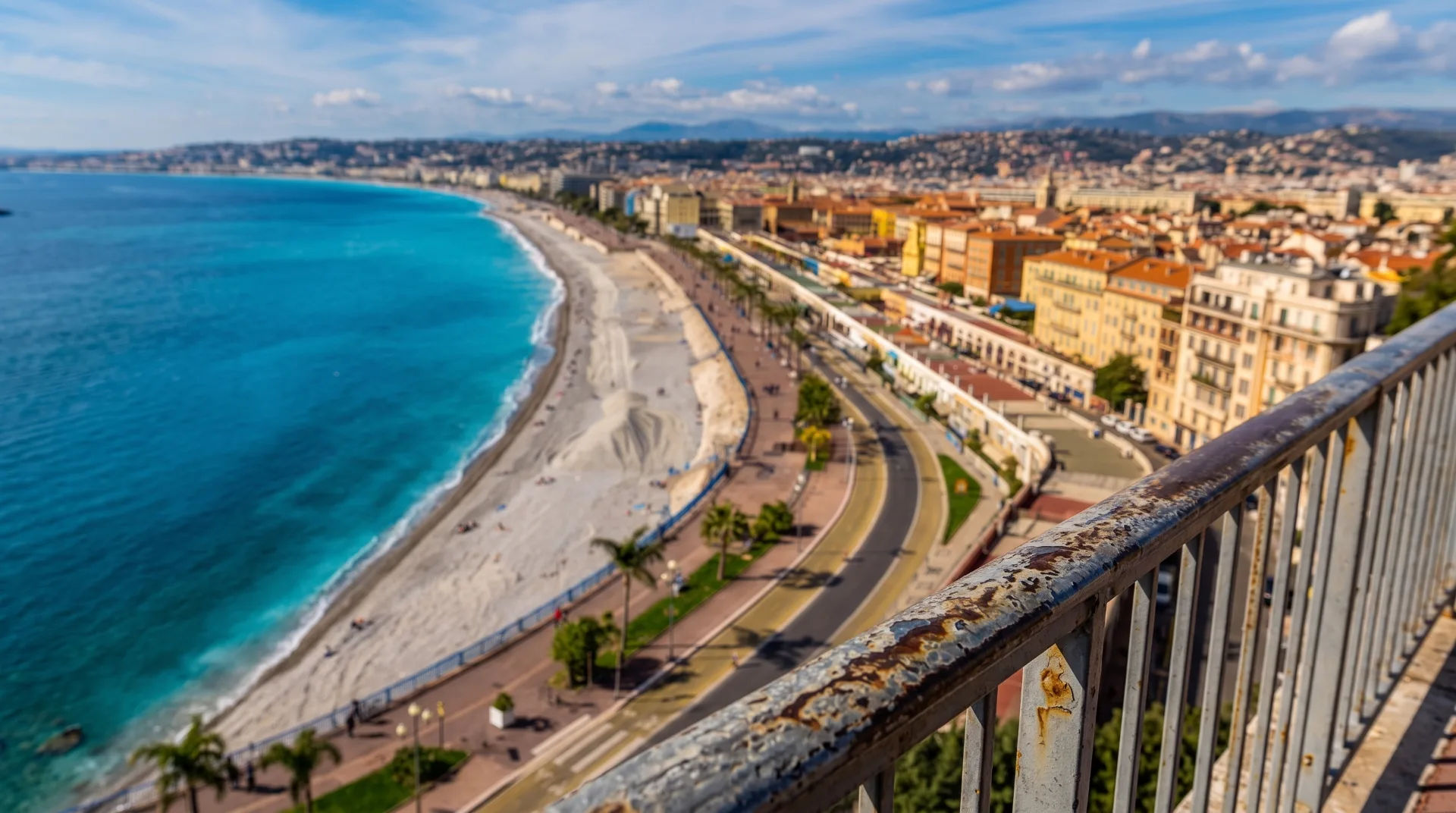 A coastal view with a sandy beach and turquoise water, lined by palm trees and buildings in the background.