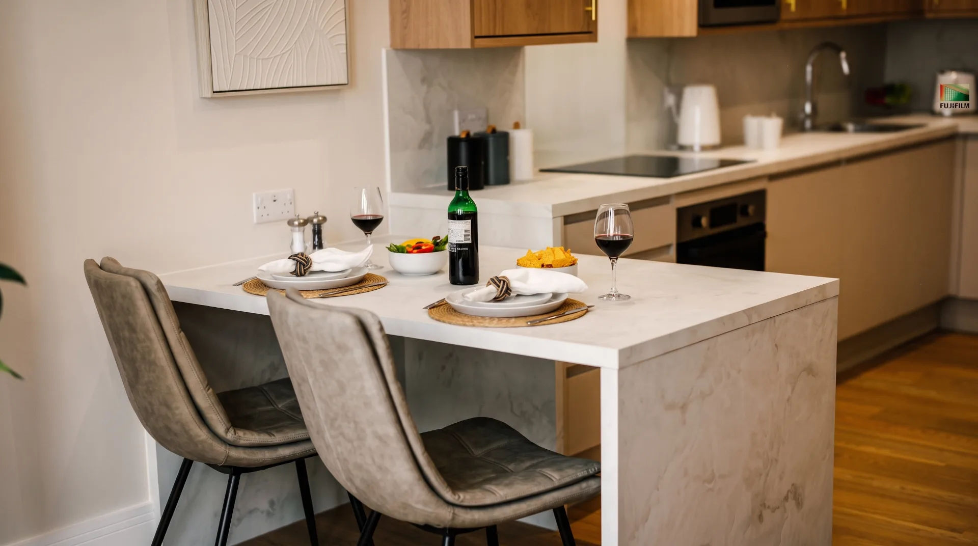 Modern kitchen area with a dining table set for two, featuring wine glasses, a bottle of wine, and a bowl of snacks.