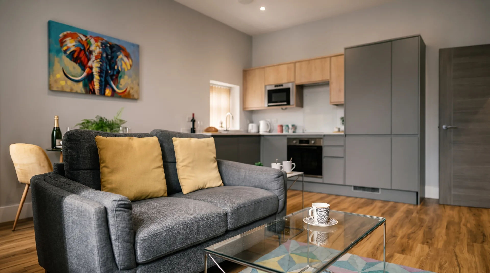 Modern living area with grey sofa, yellow cushions, glass coffee table, and kitchen in background featuring appliances.