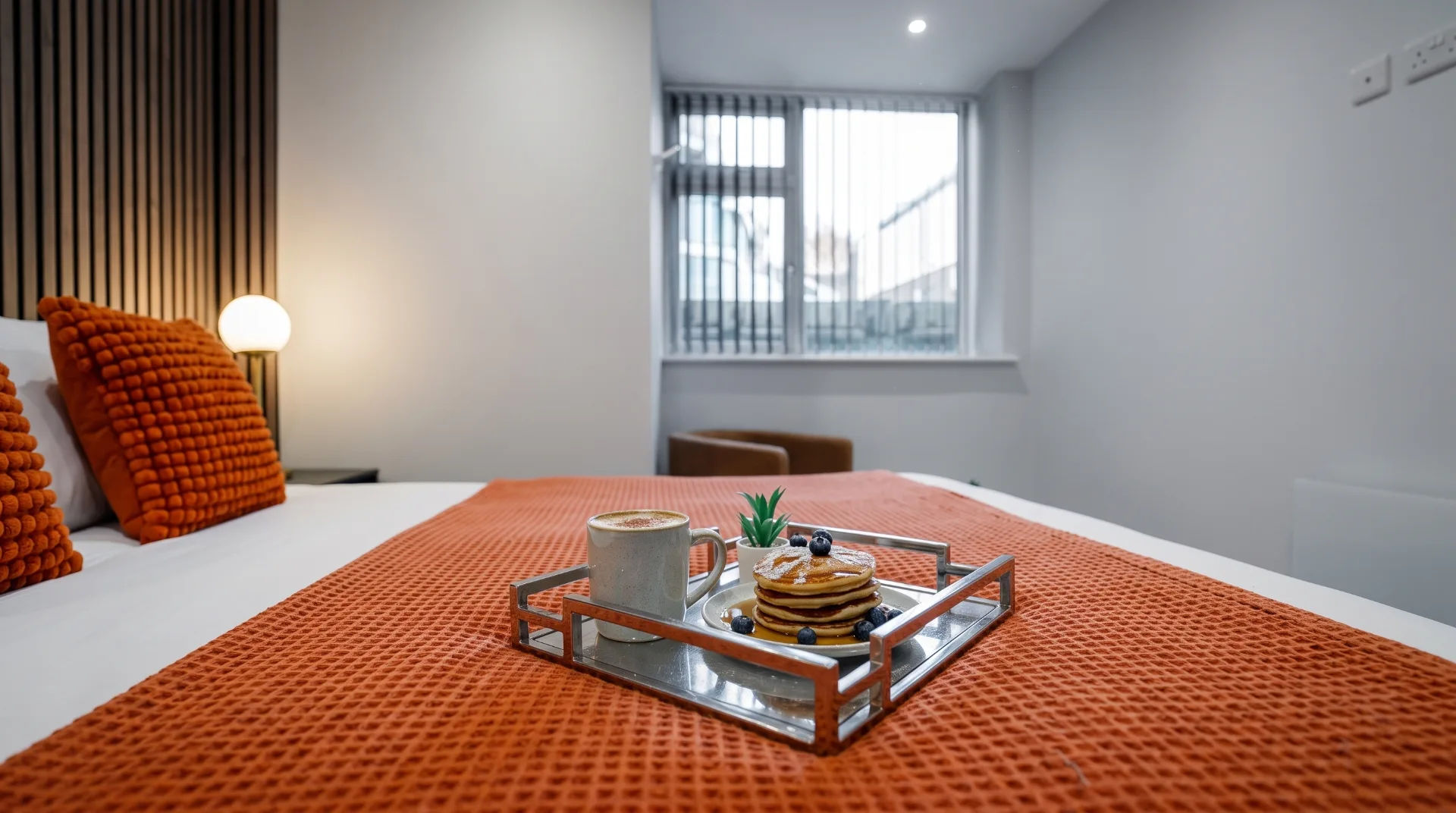 A tray with pancakes, blueberries, and a coffee cup on a bed with an orange textured throw.