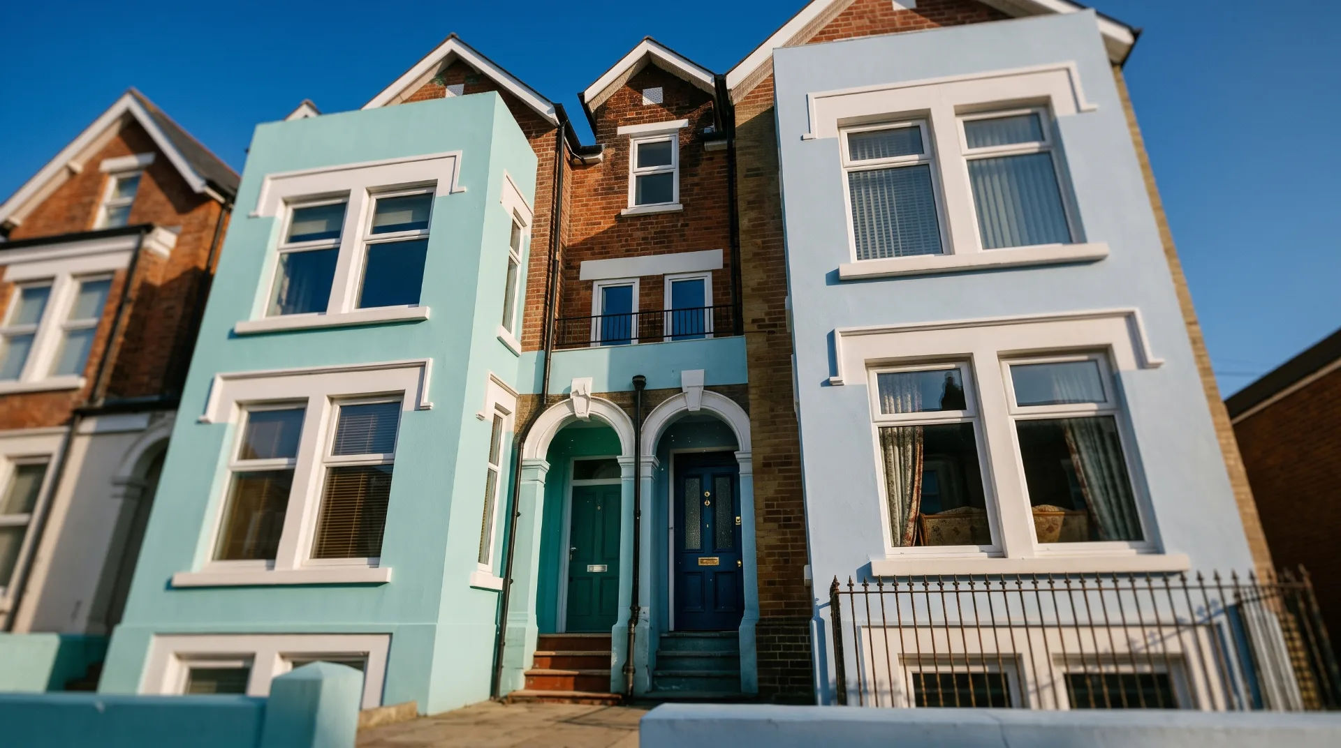 Two-storey building with turquoise and light blue facades, featuring white windows and doors, under a clear blue sky.