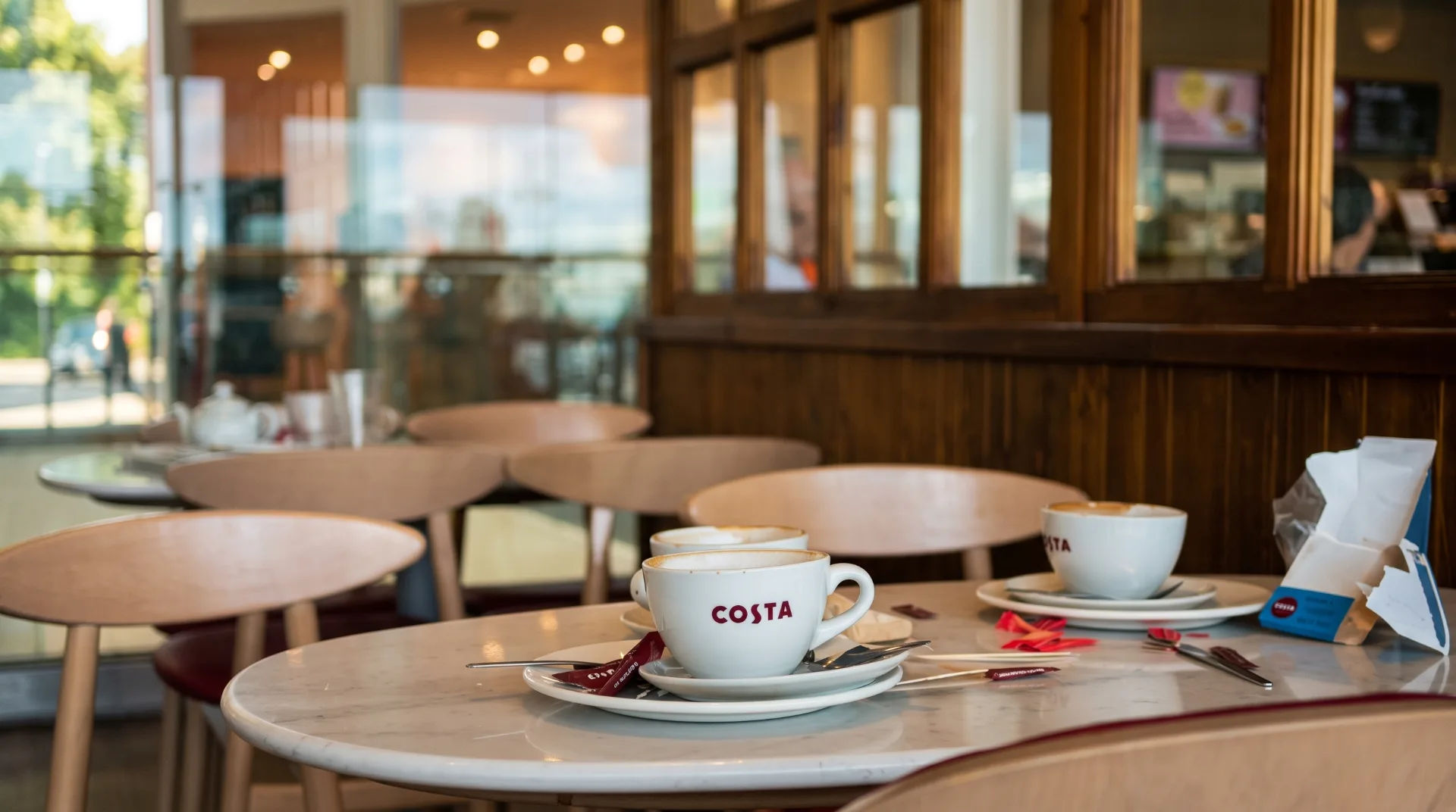 Two Costa coffee cups on a marble table with cutlery and a takeaway bag in a bright café setting.