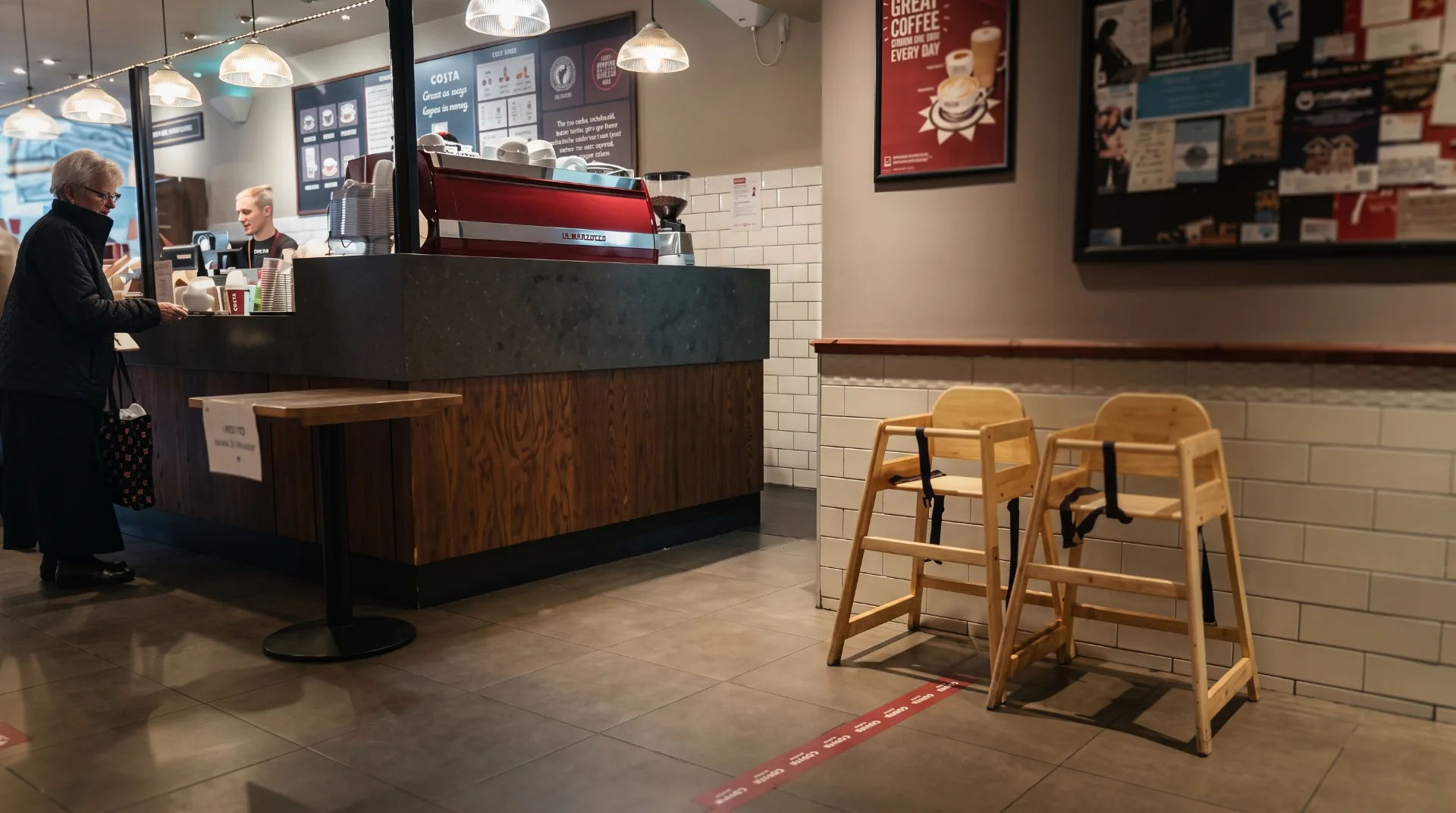 A woman at the counter of Costa with a barista behind, high chairs in the foreground and a menu on the wall