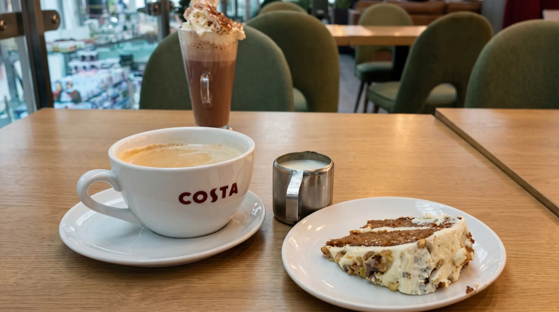 A cup of coffee, a slice of cake, and a small jug of milk on a wooden table in a Costa coffee shop.