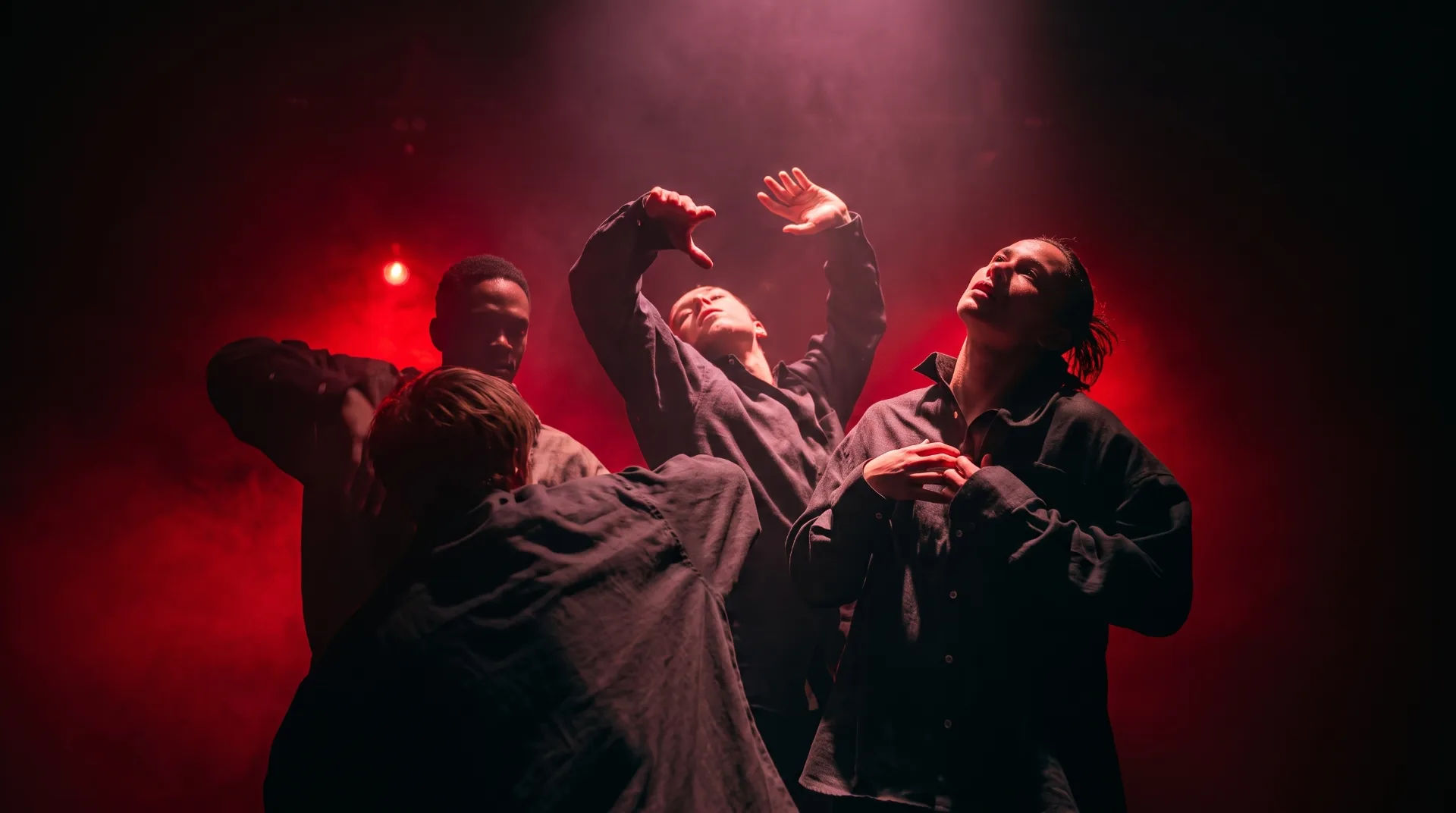 Four performers in dark shirts expressively pose against a smoky red backdrop with dramatic lighting.