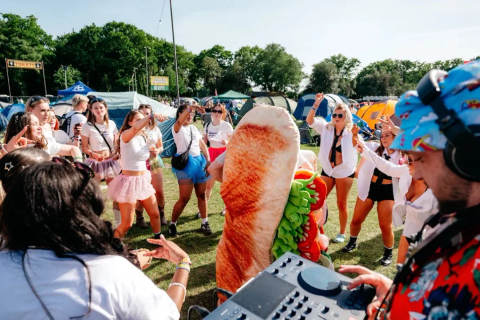 Group of festival attendees dancing near tents with a person in a food costume