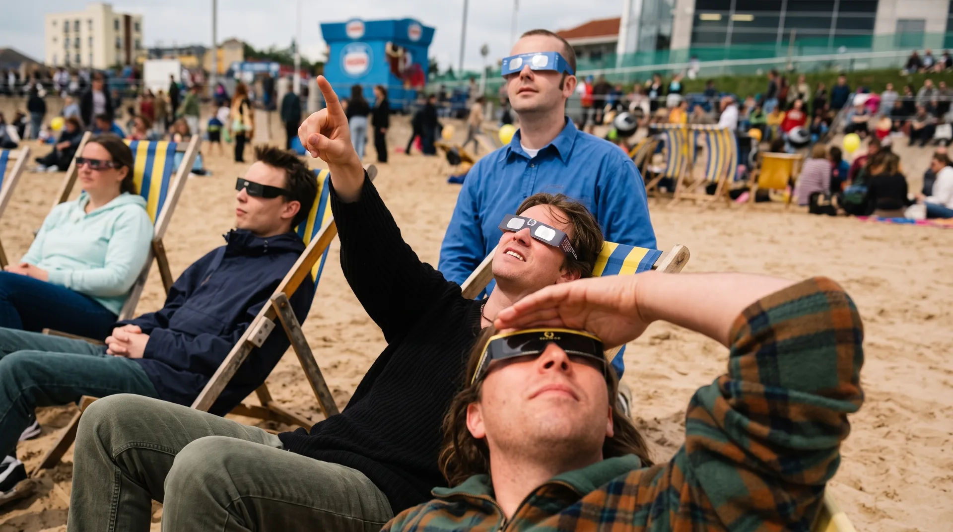 Group of people wearing eclipse glasses on the beach, looking up at the sky, with deck chairs and a crowd in the background
