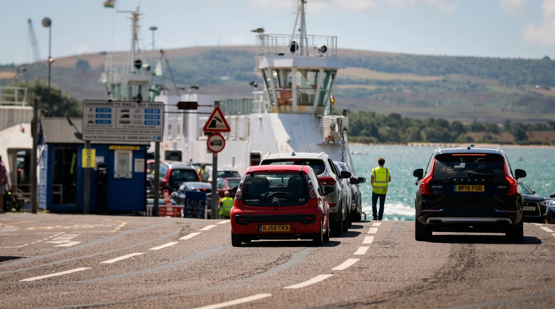 Cars lined up at Sandbanks Ferry terminal with a ferry in the background and a staff member in a high-visibility vest