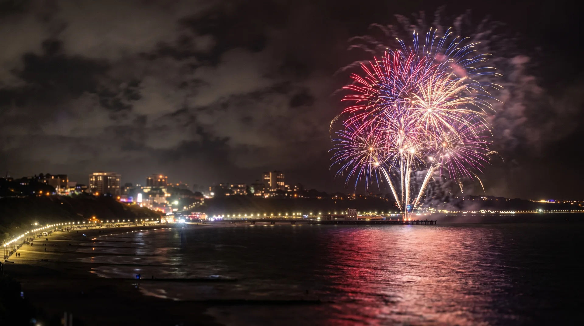 Vibrant fireworks illuminate the night sky over a coastal town, reflecting on the water below.